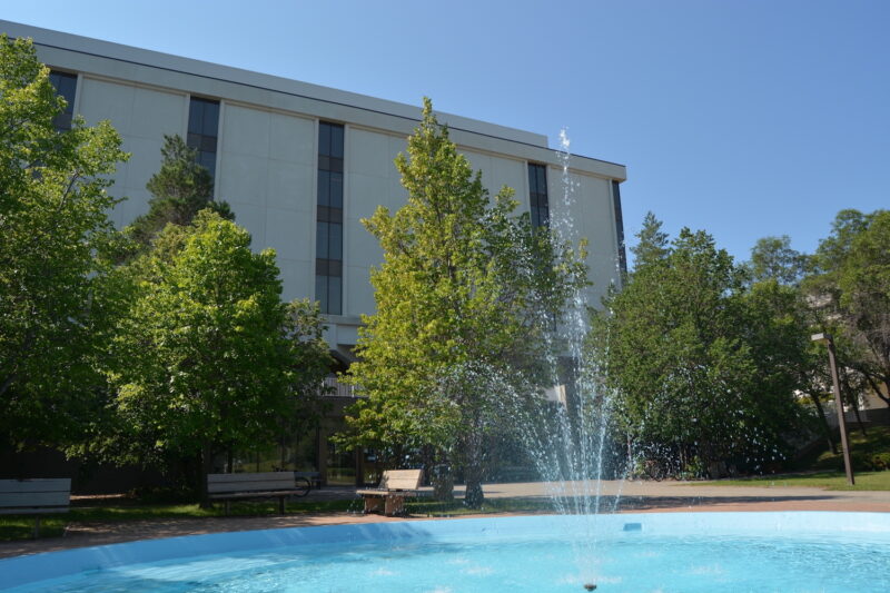 A fountain in front of the Archer Library. There are benches and trees surrounding the fountain.